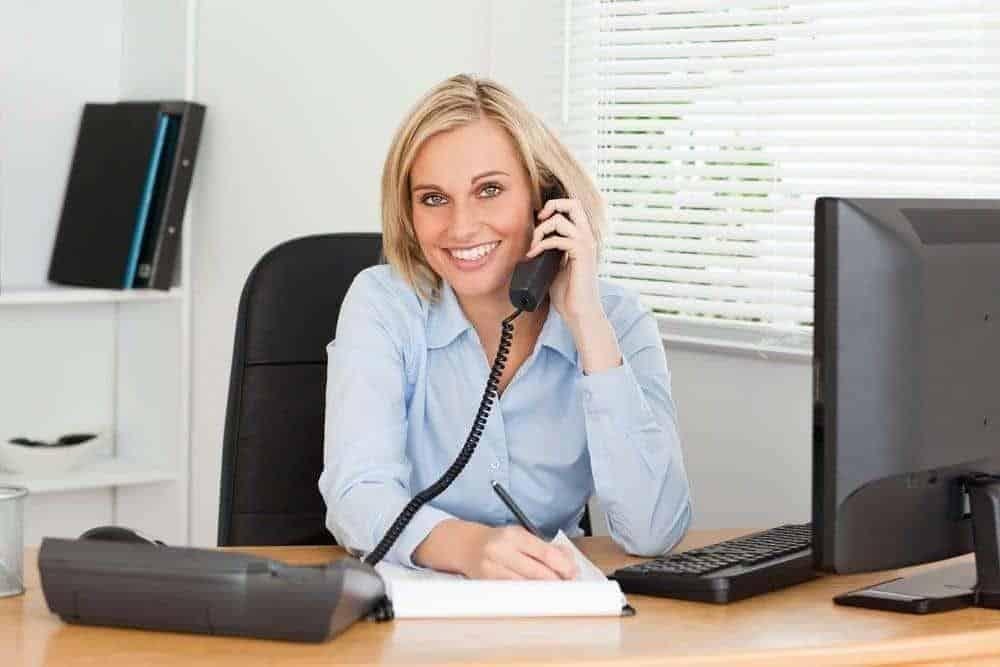 Woman in business attire talking on phone while working at office desk with computer and papers